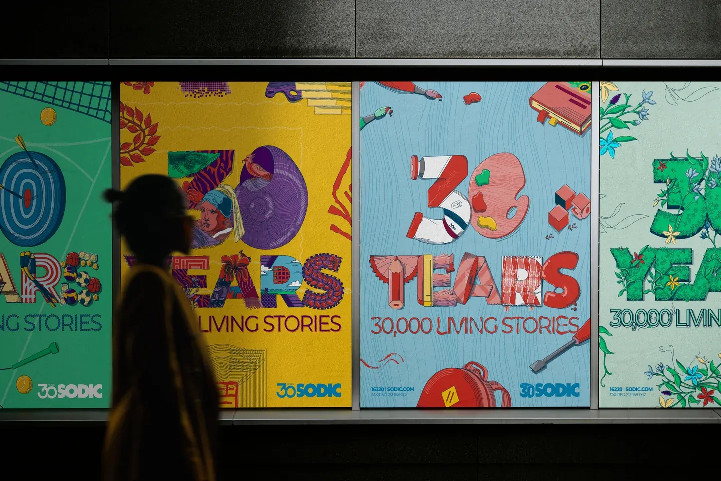 Illuminated SODIC art and education posters behind the silhouette of a passing pedestrian
