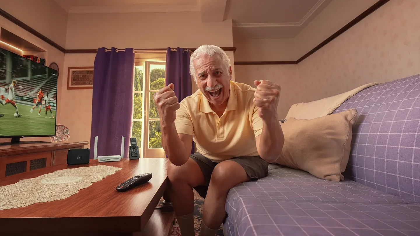 An older man cheering on a sofa beside a television showing football