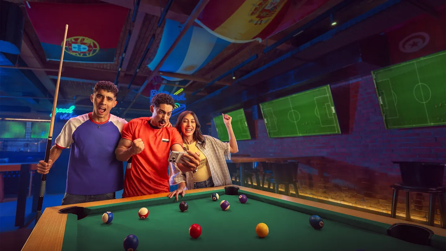 Three football fans celebrating beside a pool table in a room lit with purple and green light